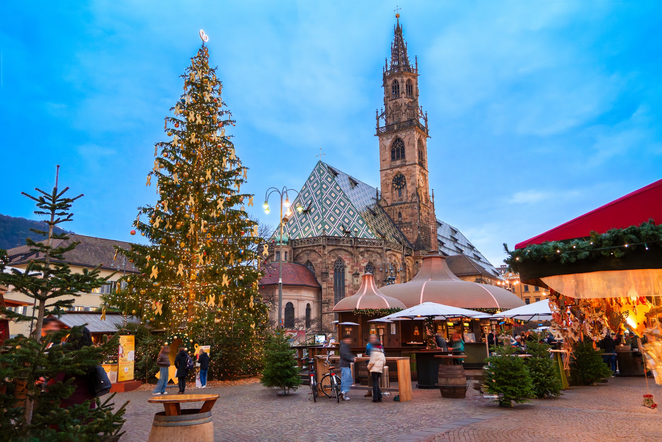 A festive Christmas market with a decorated tree, wooden stalls, and people gathered in front of a historic stone church at dusk.