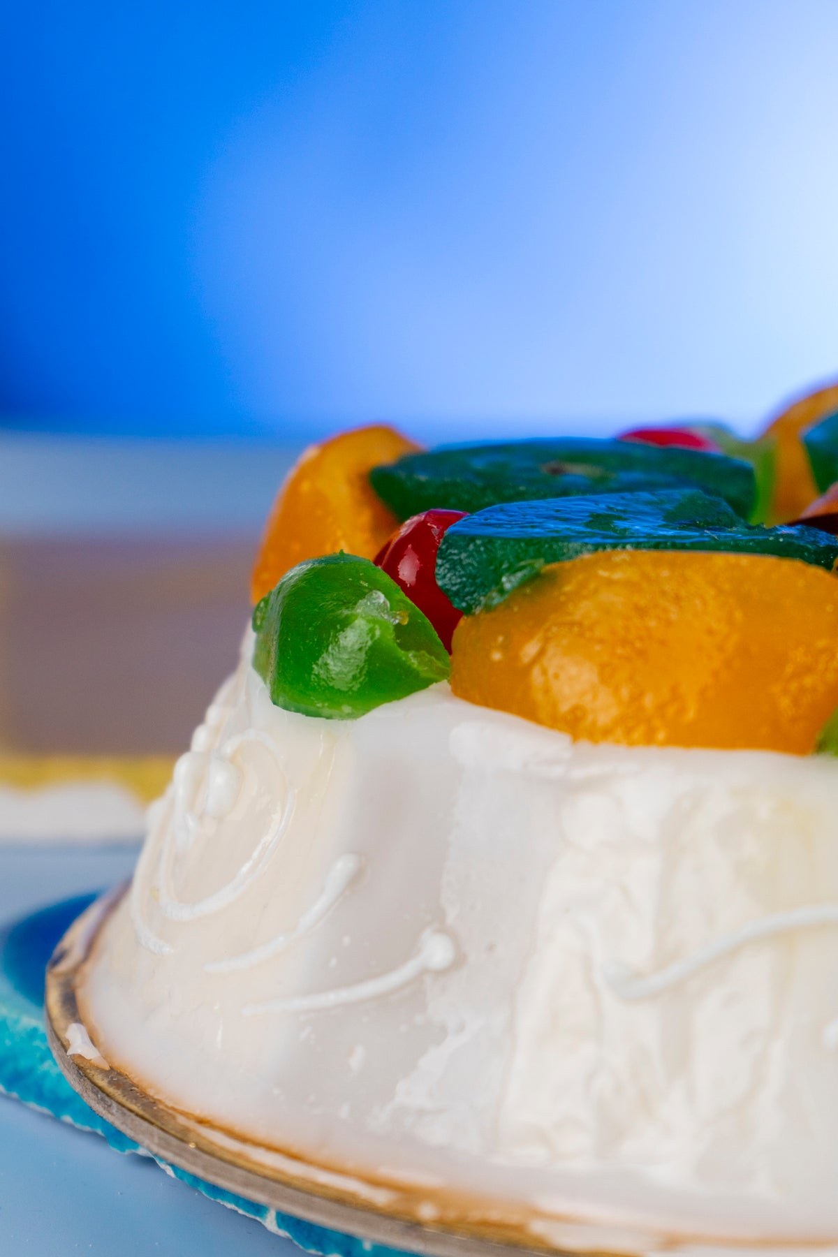 A close-up of a white frosted cake topped with colorful candied fruits, including orange, green, and red pieces, against a blue background.