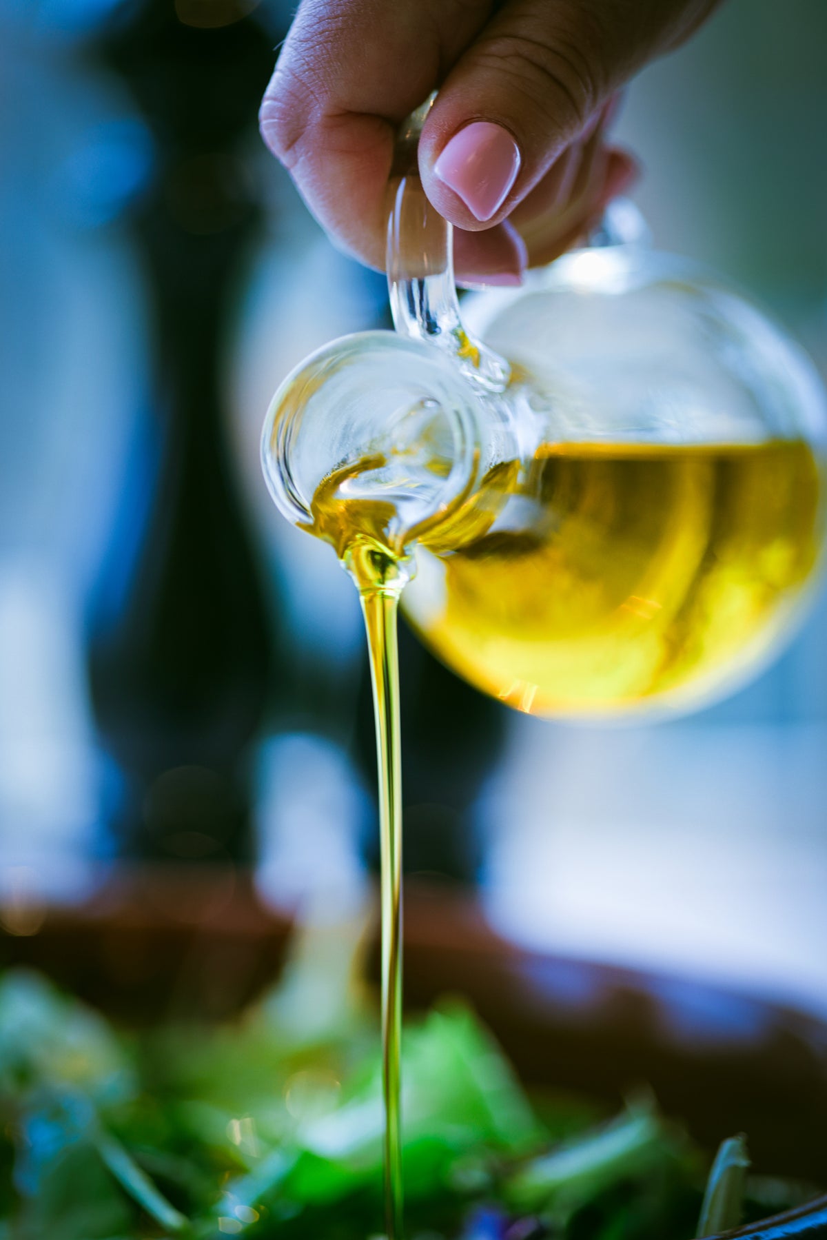A hand pours golden olive oil from a glass jug onto a fresh green salad, with a blurred background.