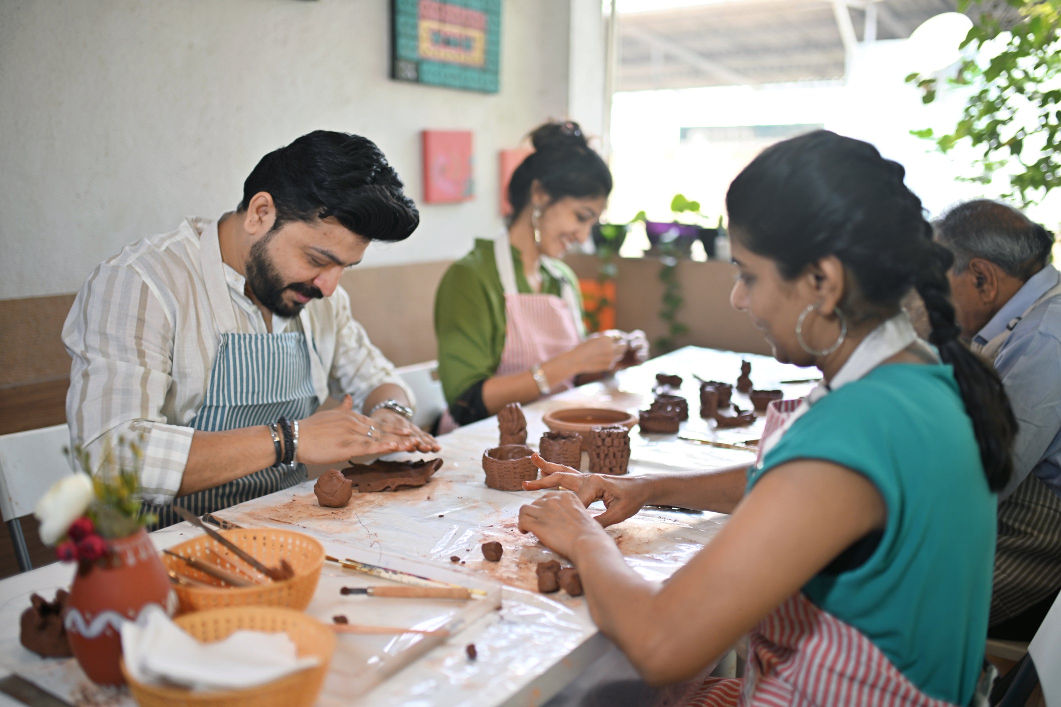 Three people sit at a table shaping clay into small sculptures, wearing aprons and focused on their work. Art supplies and finished clay pieces are scattered on the table.