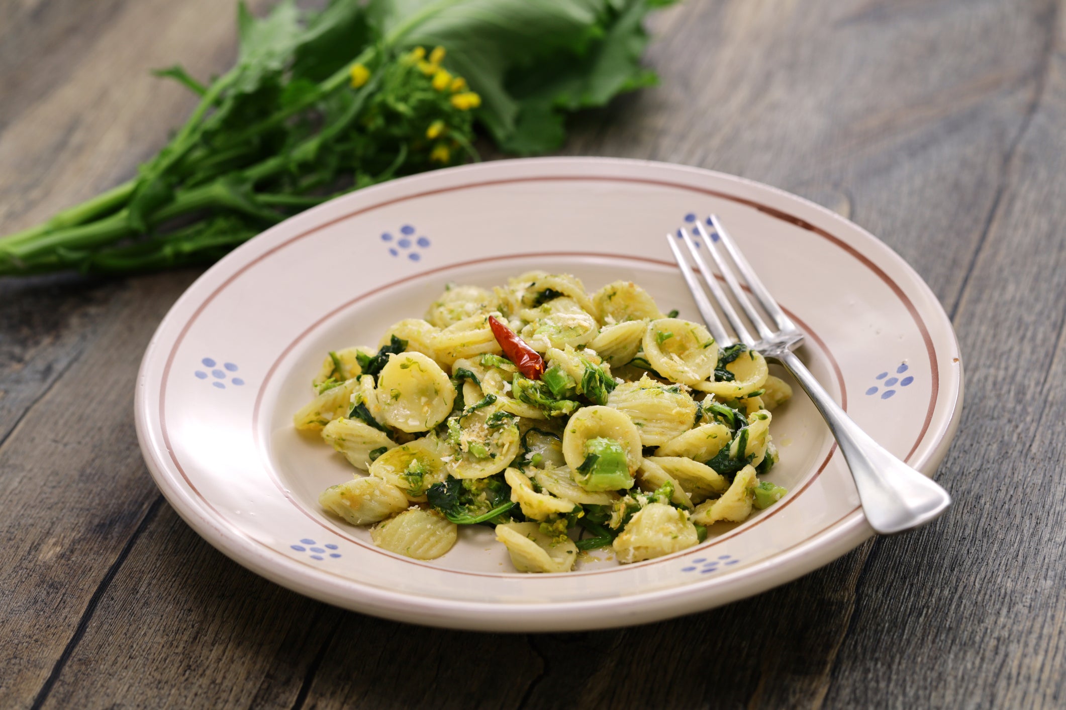 A plate of orecchiette pasta with broccoli rabe and red chili pepper, garnished with herbs, sits on a wooden table with a fork and fresh greens in the background.