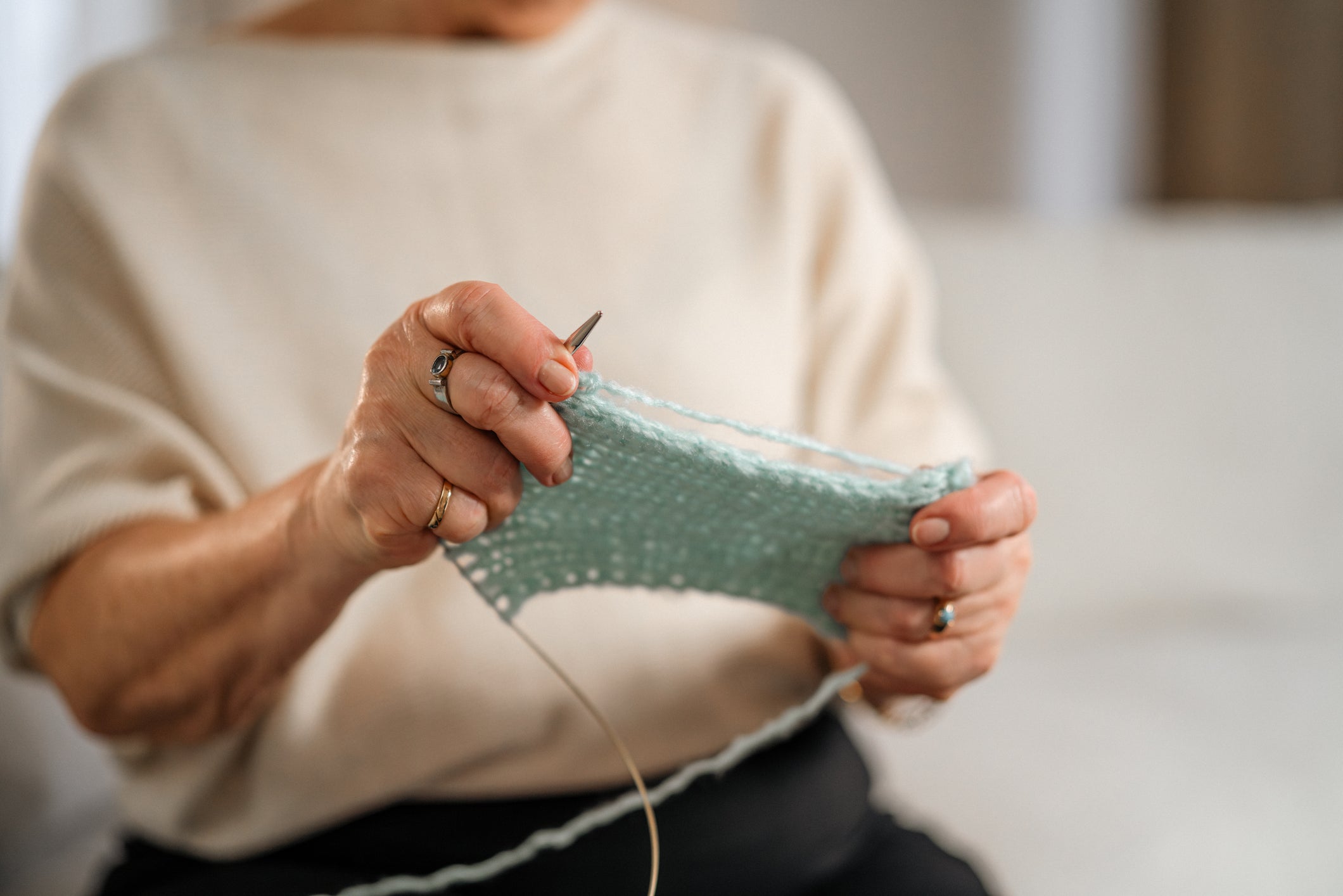 A person wearing a beige sweater is knitting with light blue yarn, holding knitting needles and working on a textured fabric piece.