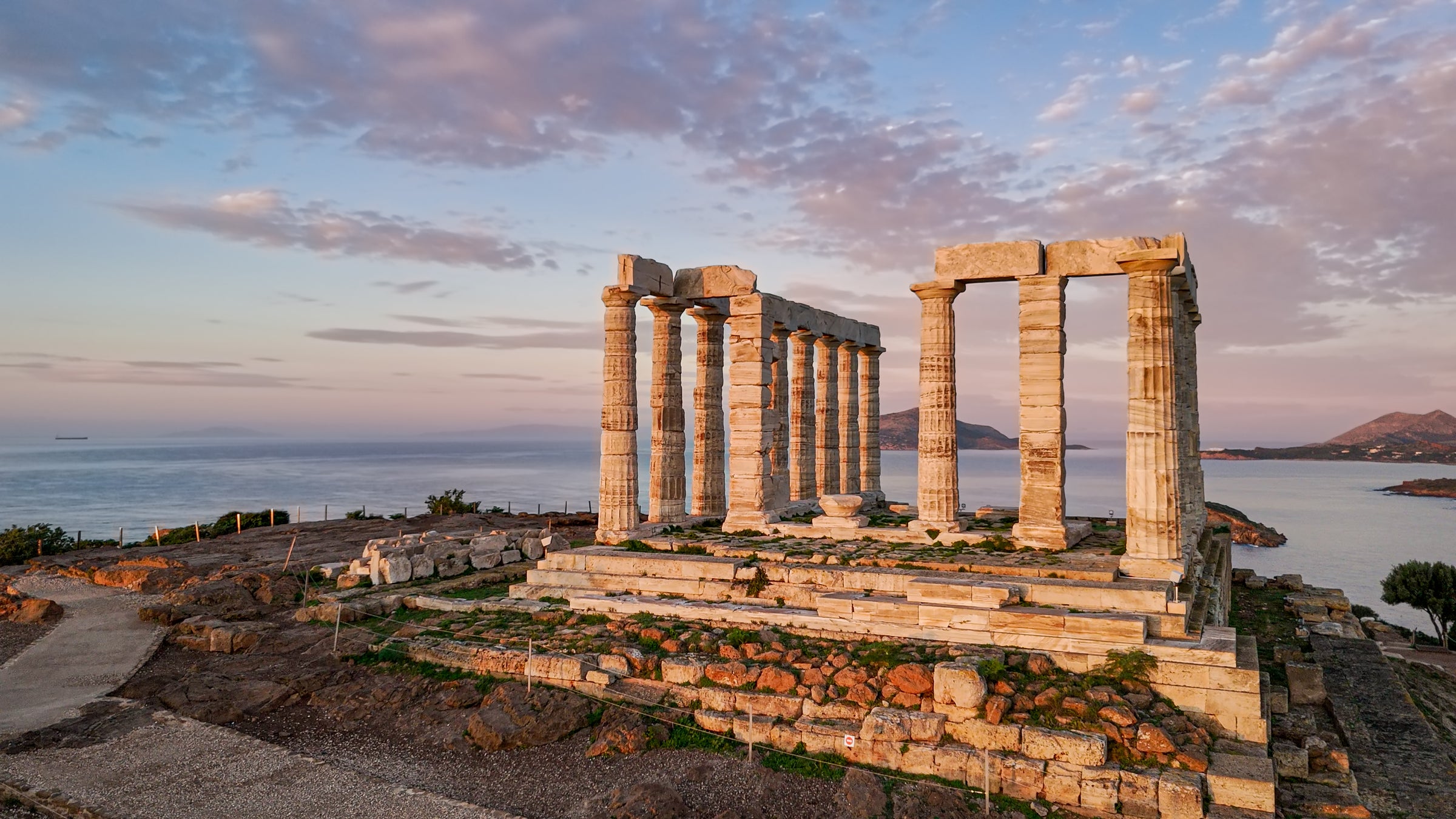 Ancient Greek temple ruins with tall stone columns stand on a rocky hill overlooking the sea at sunset, with soft clouds and distant land visible in the background.