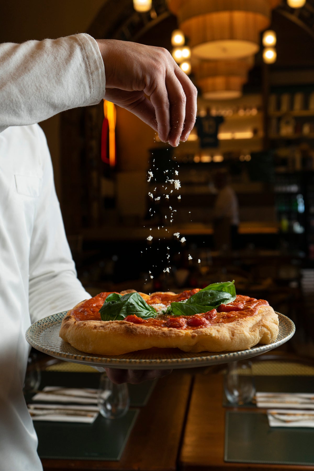 A person sprinkles grated cheese over a freshly baked pizza topped with basil leaves, served on a plate in a warmly lit restaurant.