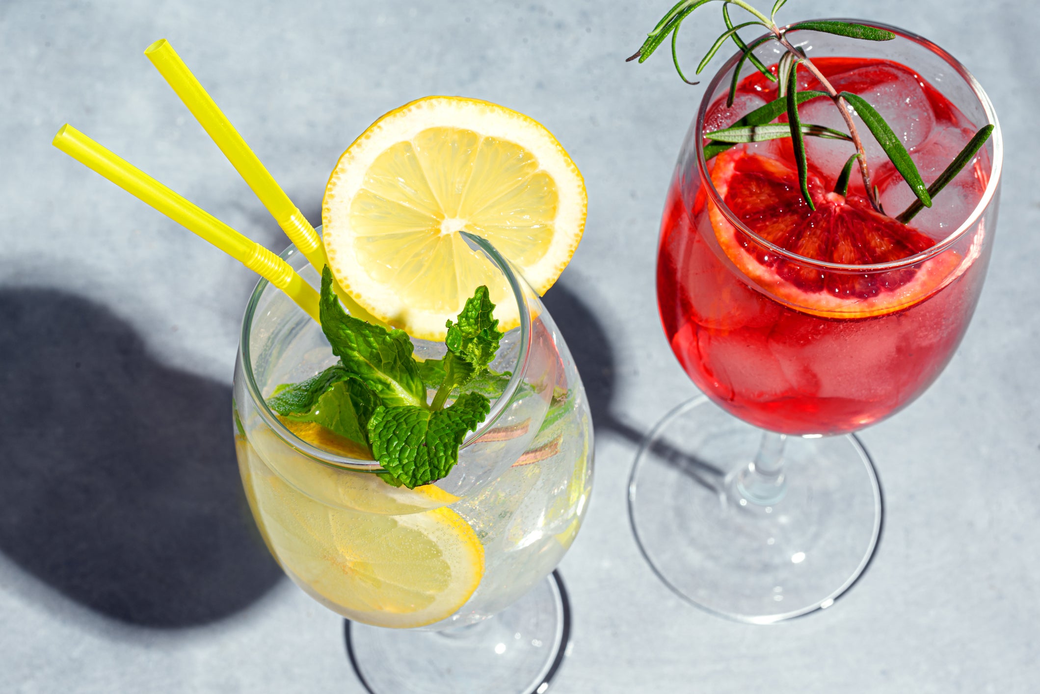 Two glasses of drinks: one with lemon slices, mint, yellow straws, and ice; the other with red liquid, blood orange slices, rosemary, and ice. Both are on a light gray surface with strong sunlight and shadows.