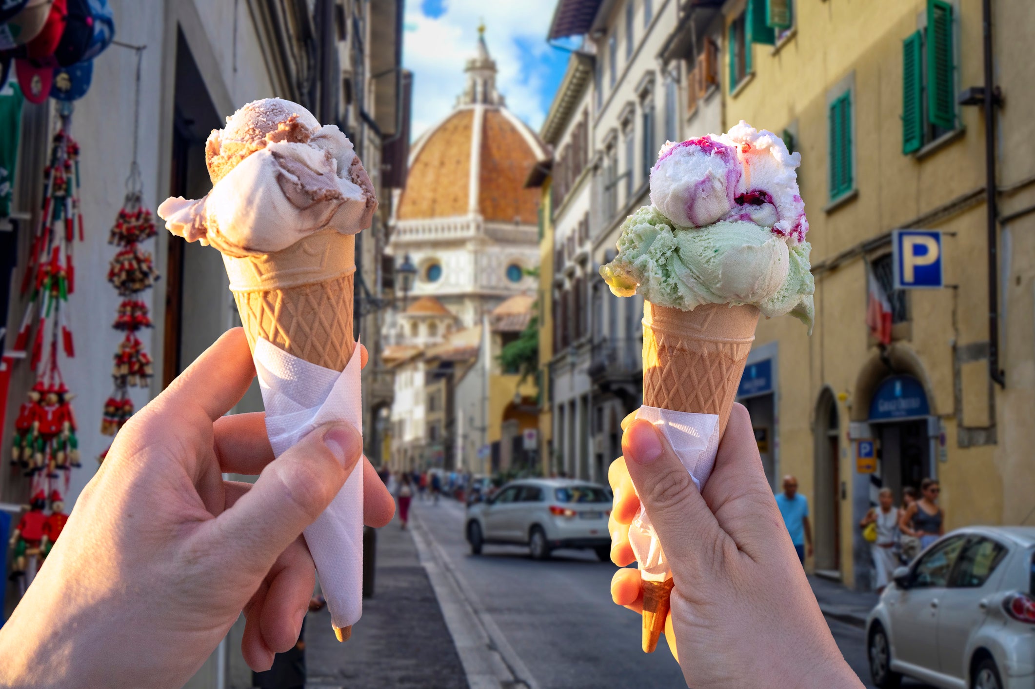 Two hands hold ice cream cones on a street in Florence, Italy, with the Cathedral of Santa Maria del Fiore (Duomo) visible in the background and cars parked along the buildings.