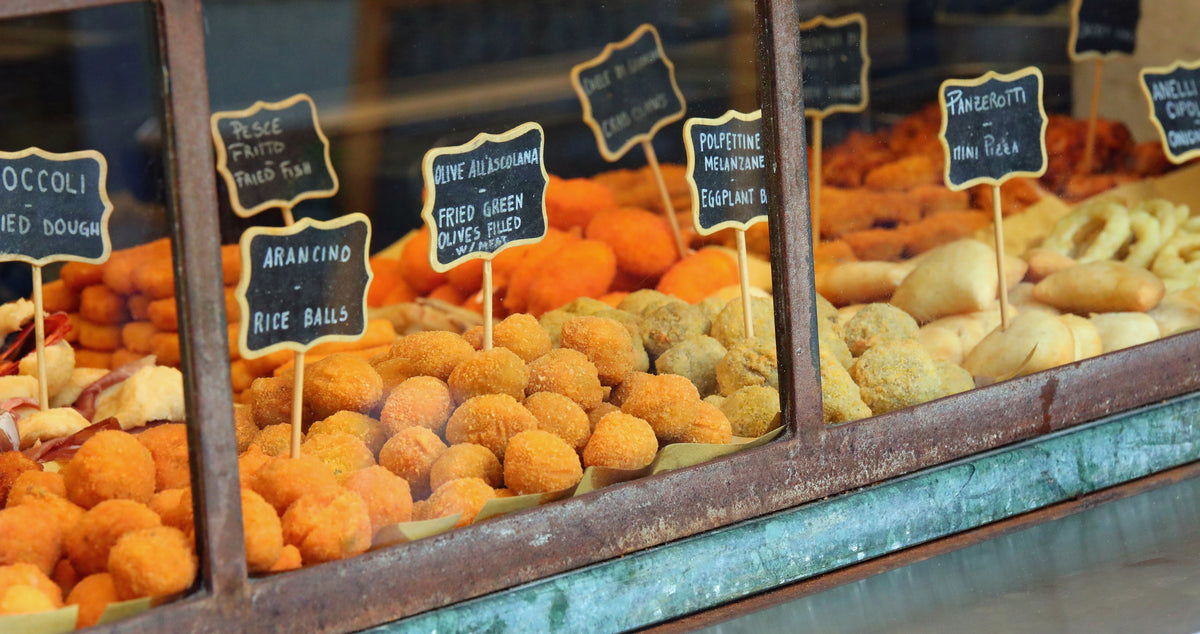 A display case filled with various fried Italian street foods, each item labeled with small blackboard signs, including arancini, panzerotti, and other golden, breaded snacks.