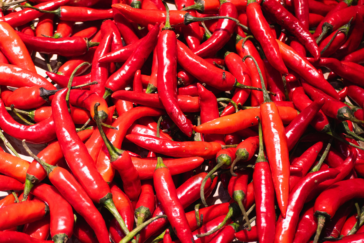 A pile of fresh red chili peppers, some with green stems, filling the entire frame. The peppers are glossy and vary in size, laying in different directions.