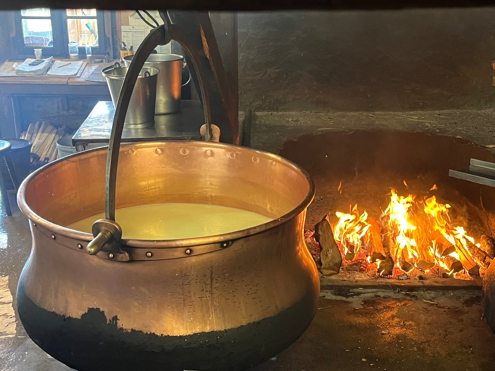 A large copper cauldron with liquid inside hangs over a wood fire in a rustic kitchen, with wooden shelves, buckets, and tools in the background.