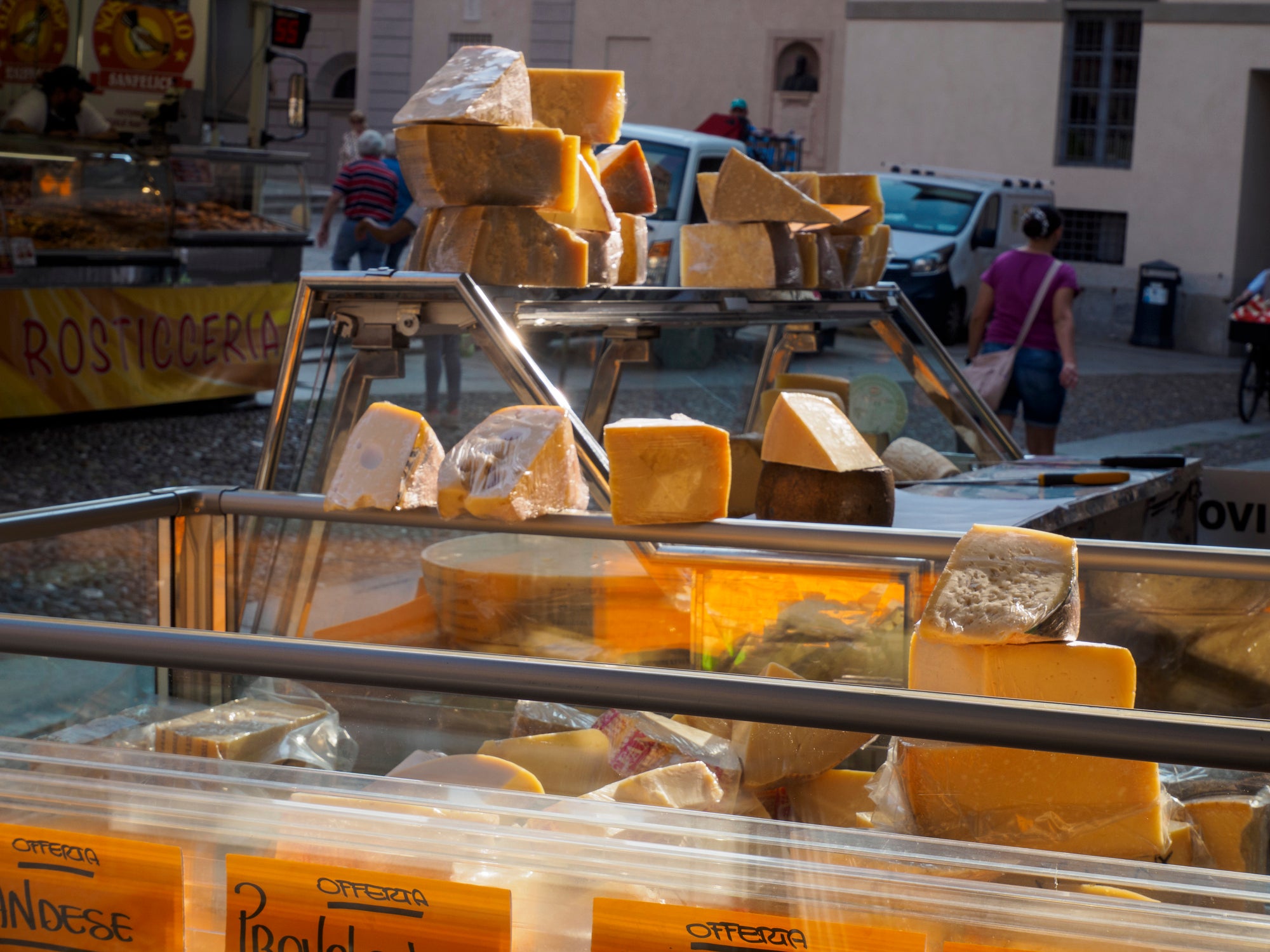 A market stall displays various large blocks and wedges of cheese in a glass case, with people and buildings visible in the background.