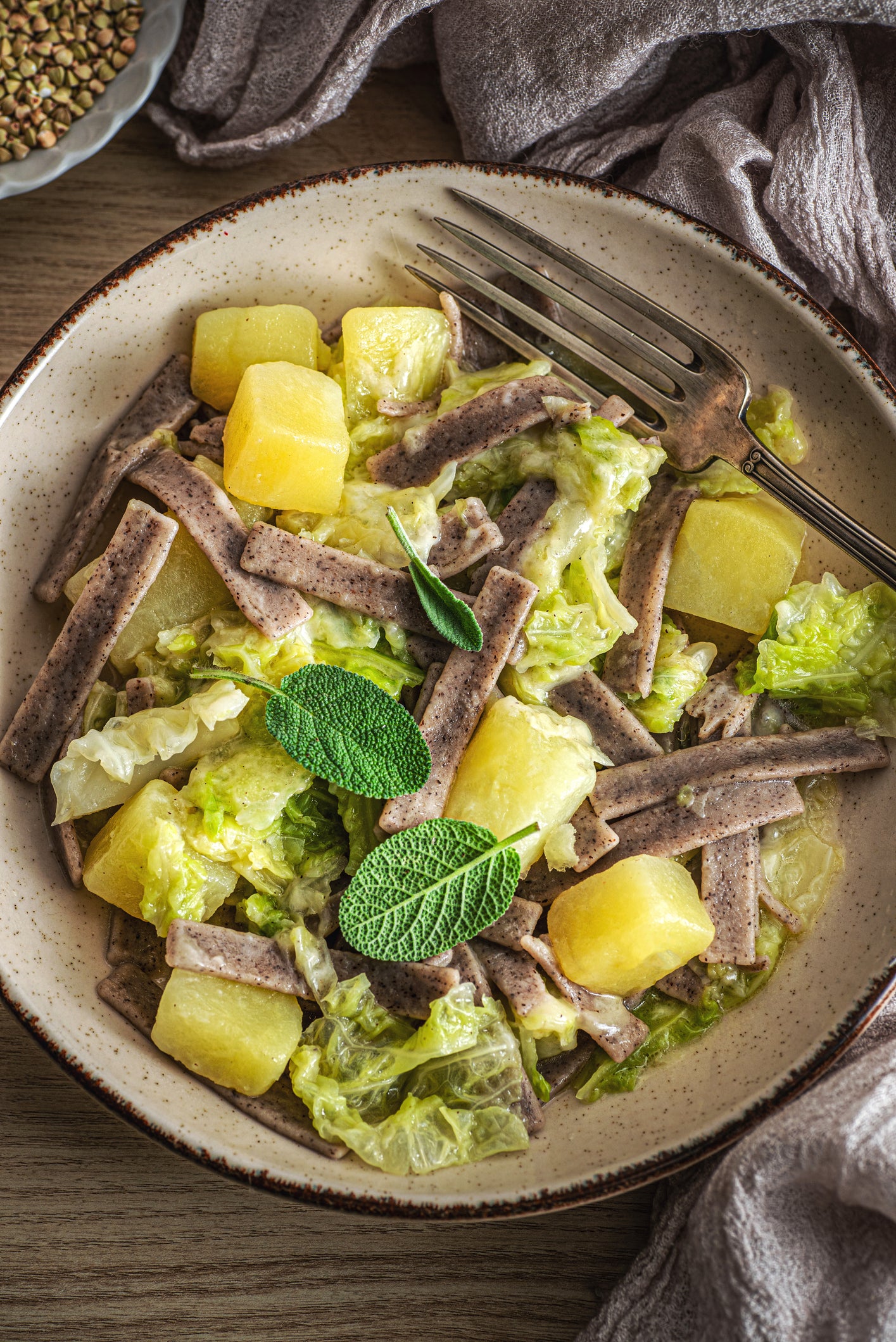 A bowl of pasta with yellow potato cubes, green cabbage pieces, and a creamy sauce, garnished with three fresh sage leaves, sits on a wooden table with a fork resting on the side.