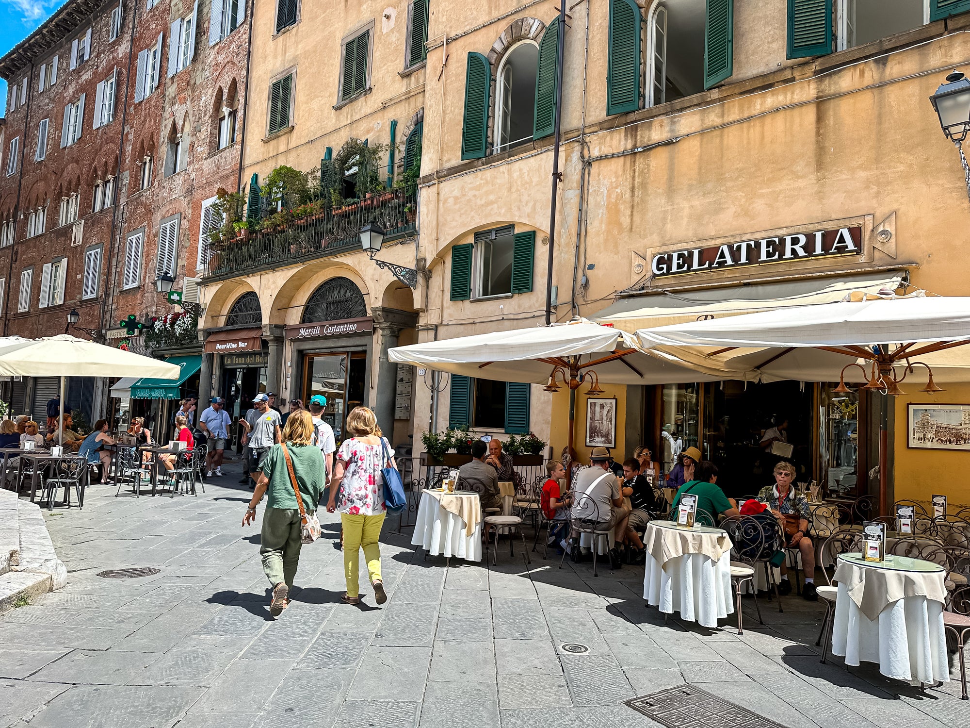 People walk along a sunny cobblestone street in Italy, passing outdoor tables at a gelateria and other shops, with colorful old buildings and green window shutters in the background.