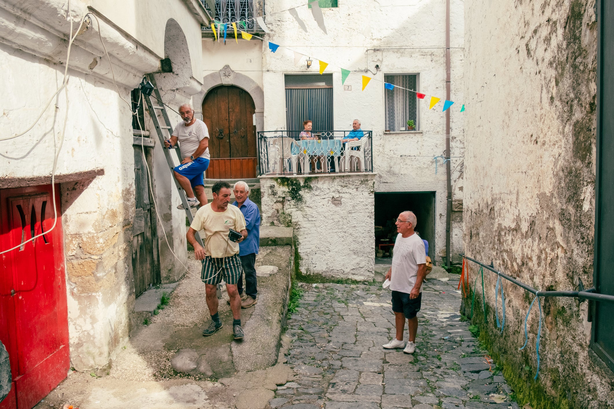 In a rustic alley, men help another man climb through a window as others watch. Colorful flags hang overhead, and several people sit on a balcony above the scene.