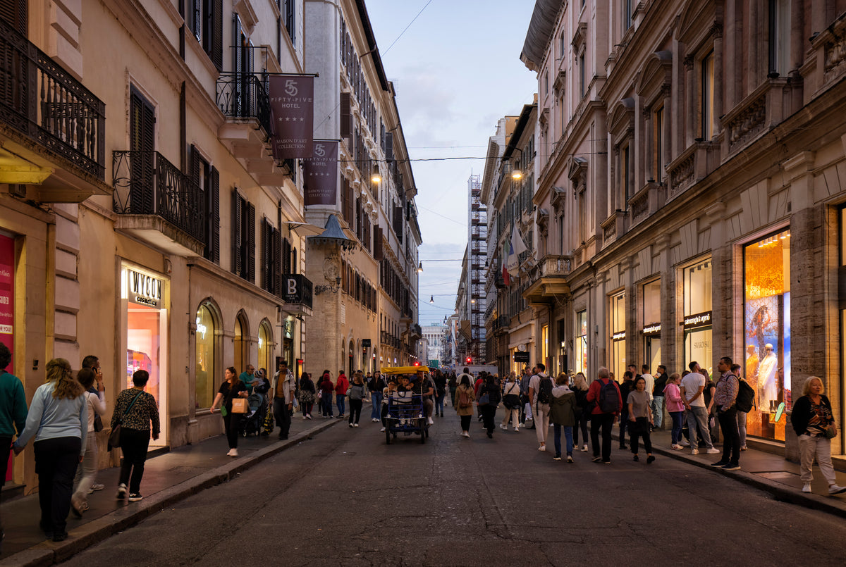 Crowds walk along a lively, narrow city street lined with shops and historic buildings at dusk, under a cloudy sky.