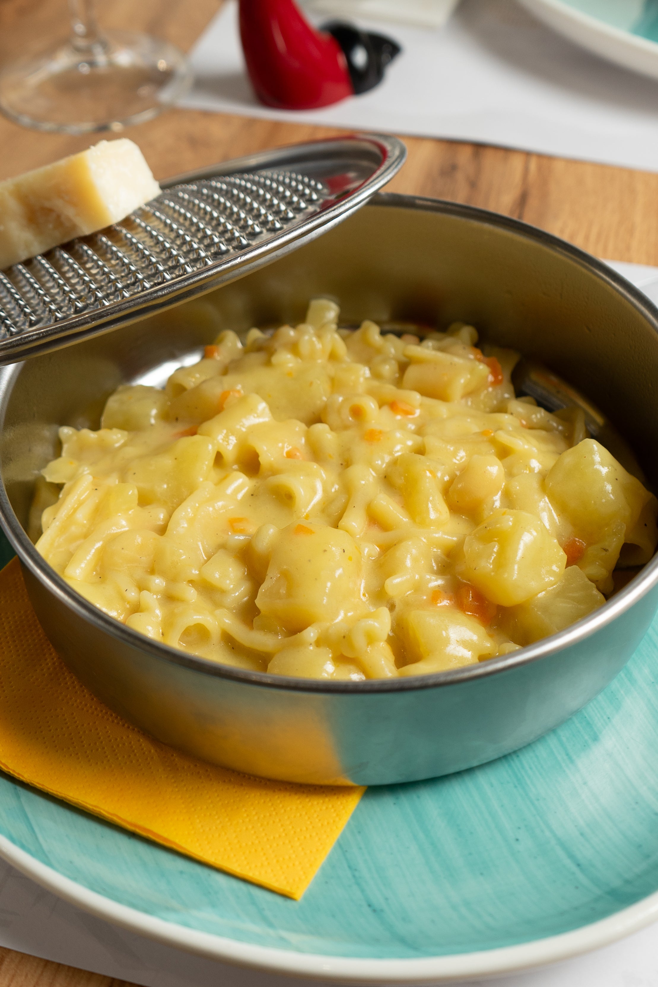 A metal bowl filled with creamy macaroni and cheese sits on a blue plate with a yellow napkin. A cheese grater with a wedge of cheese rests on the bowls rim.