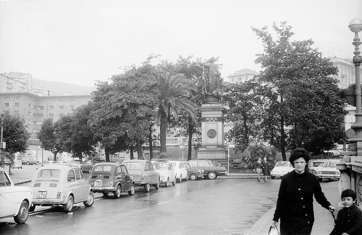 Black-and-white photo of a city street with parked vintage cars, trees, a statue on a pedestal, and two people, an adult and a child, walking in the foreground on a rainy day.