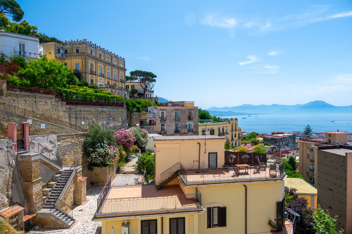 Colorful buildings on a hillside overlook the sea under a clear blue sky, with lush greenery, blooming trees, and distant mountains visible across the water.