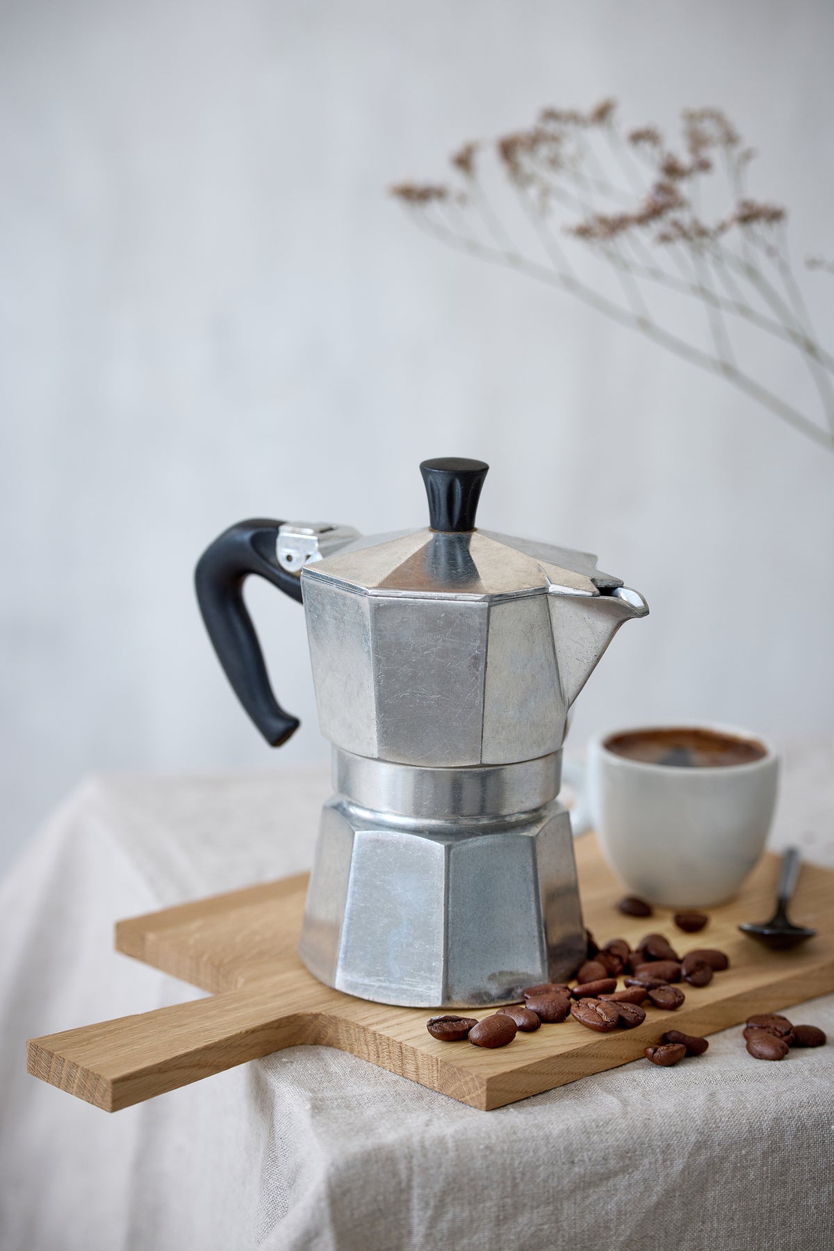 A silver moka pot sits on a wooden board with scattered coffee beans, next to a white cup of coffee and a spoon. Dried flowers are blurred in the background.