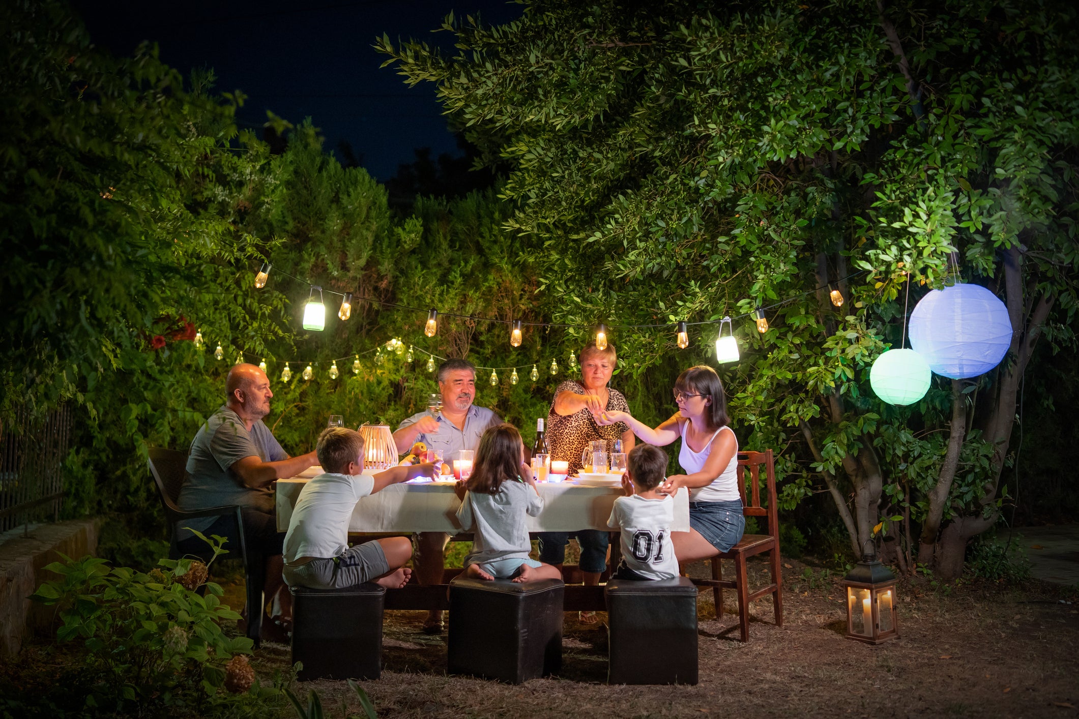 Six people, including two children, sit around a table outdoors at night, enjoying a meal under string lights and paper lanterns. The garden is lush with greenery, creating a warm, festive atmosphere.