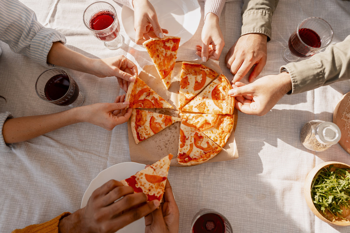 Five people reach for slices of cheese pizza on a table set with glasses of red beverage, a salad, and a light tablecloth, creating a casual and inviting dining scene.