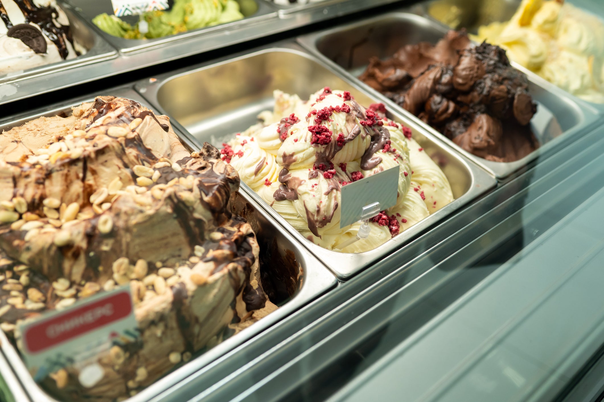 Assorted gelato flavors in metal trays inside a display case, including chocolate with nuts, vanilla with raspberry pieces, and rich chocolate, viewed from above.