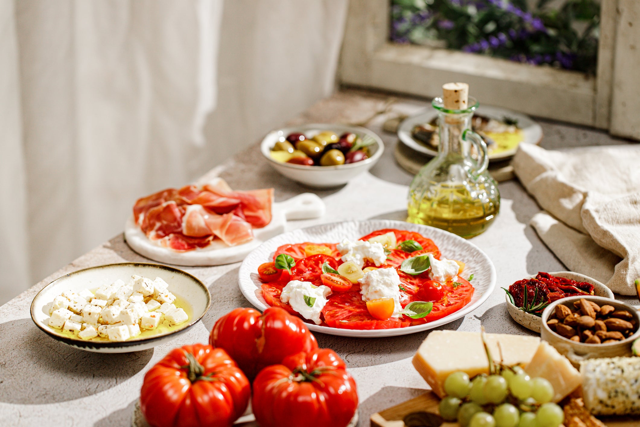 A table with sliced tomatoes and cheese, fresh basil, prosciutto, olives, olive oil, feta, nuts, grapes, and cheese, all set in natural sunlight.