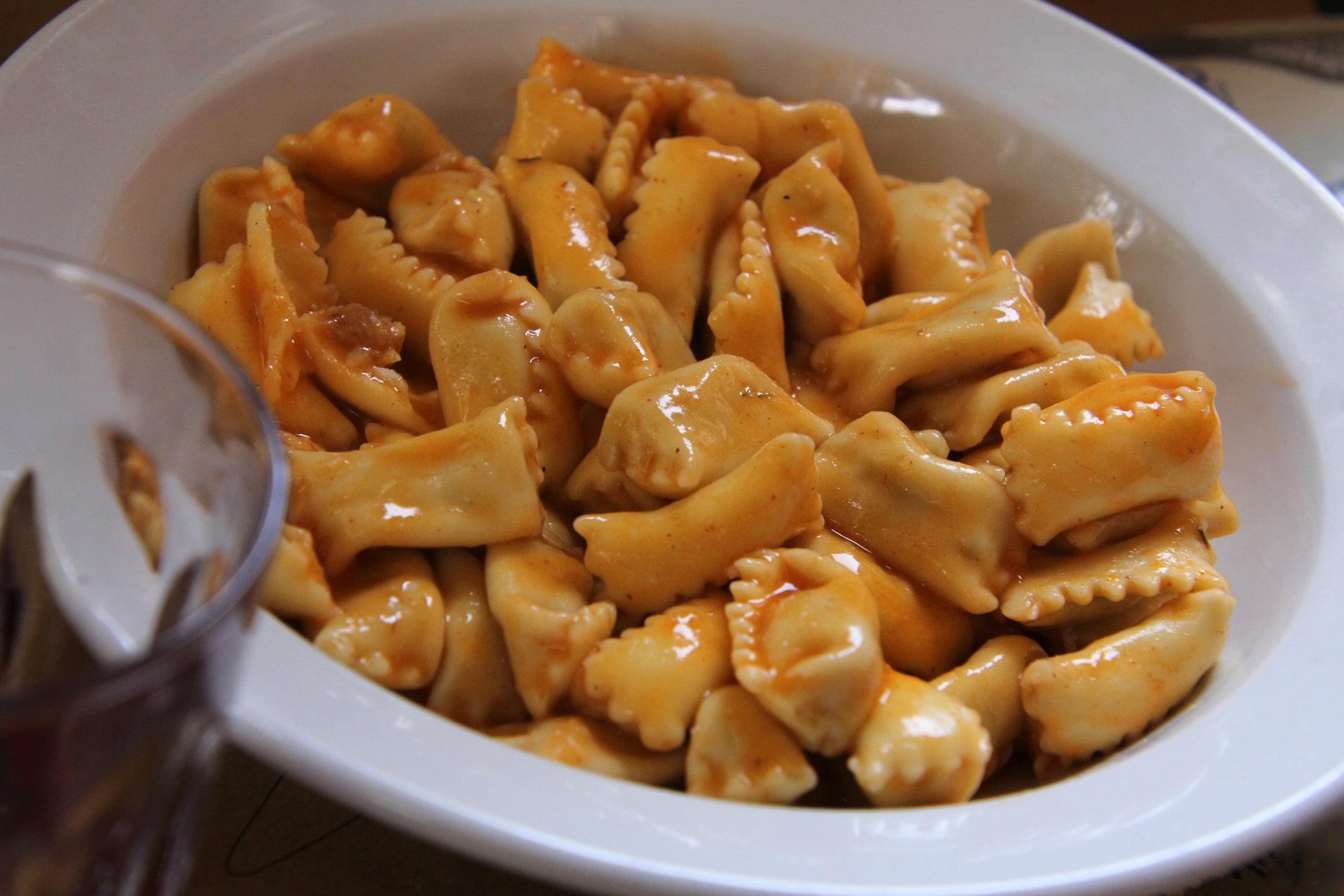 A white bowl filled with small, square-shaped pasta pieces coated in a light tomato sauce. Part of a glass is visible in the foreground.