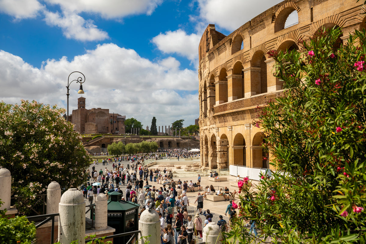 The Colosseum in Rome on a sunny day, surrounded by greenery and blooming flowers, with many tourists gathered outside and ancient ruins visible in the background.