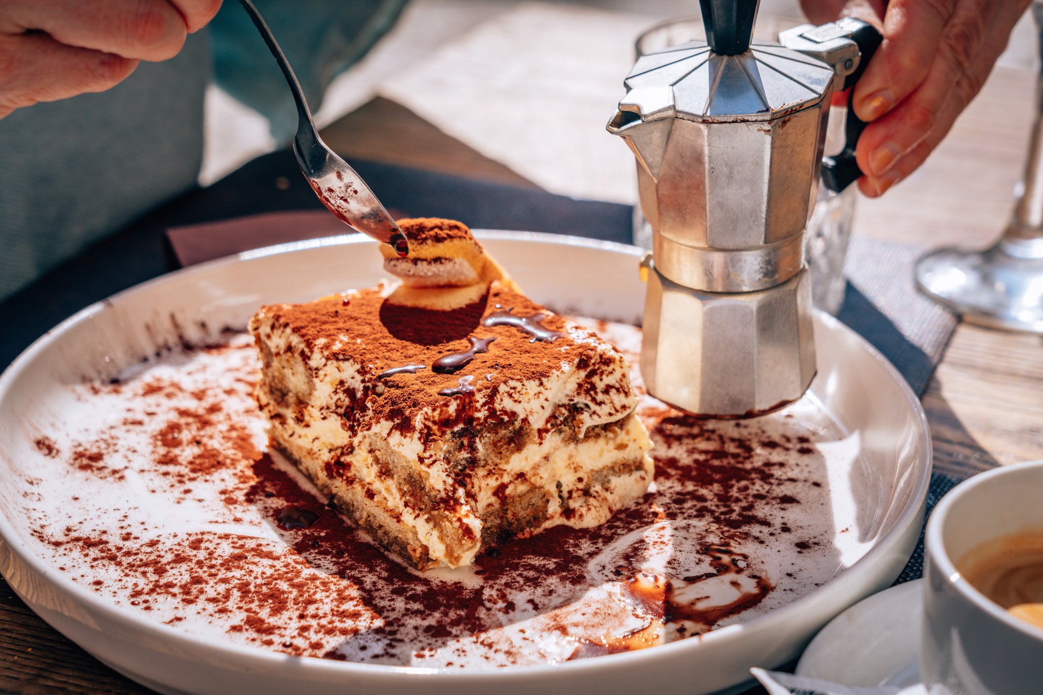 A close-up of a hand holding a fork about to eat tiramisu dusted with cocoa powder, with a small moka pot and a cup of coffee in the background on a sunlit table.