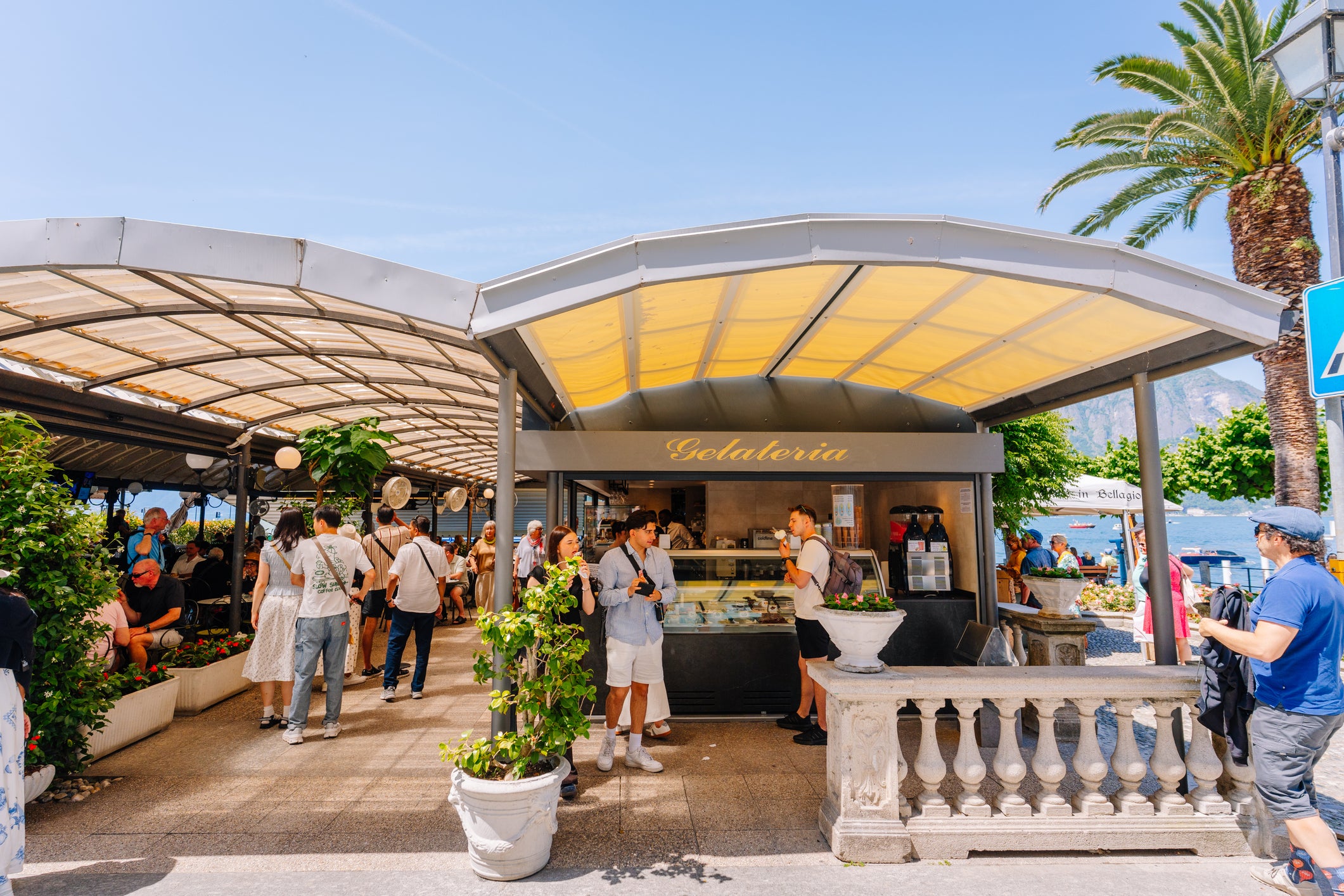 People gather around an outdoor gelateria by a lake, with palm trees, mountains, and clear blue skies in the background. Some people are ordering ice cream, while others walk or chat nearby.