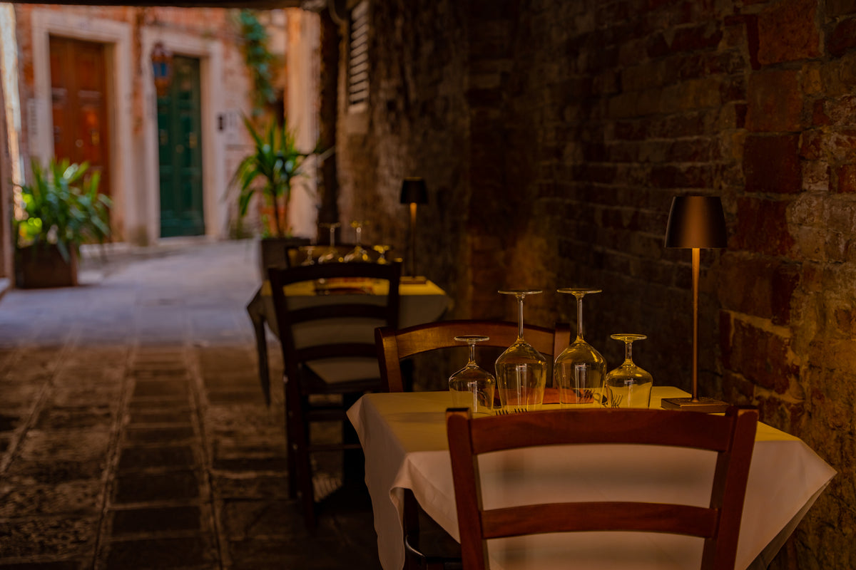 A narrow alleyway with tables set for dining, covered in yellow cloths and wine glasses, next to rustic brick walls. Soft light creates a cozy, intimate atmosphere, with plants and a doorway visible in the background.