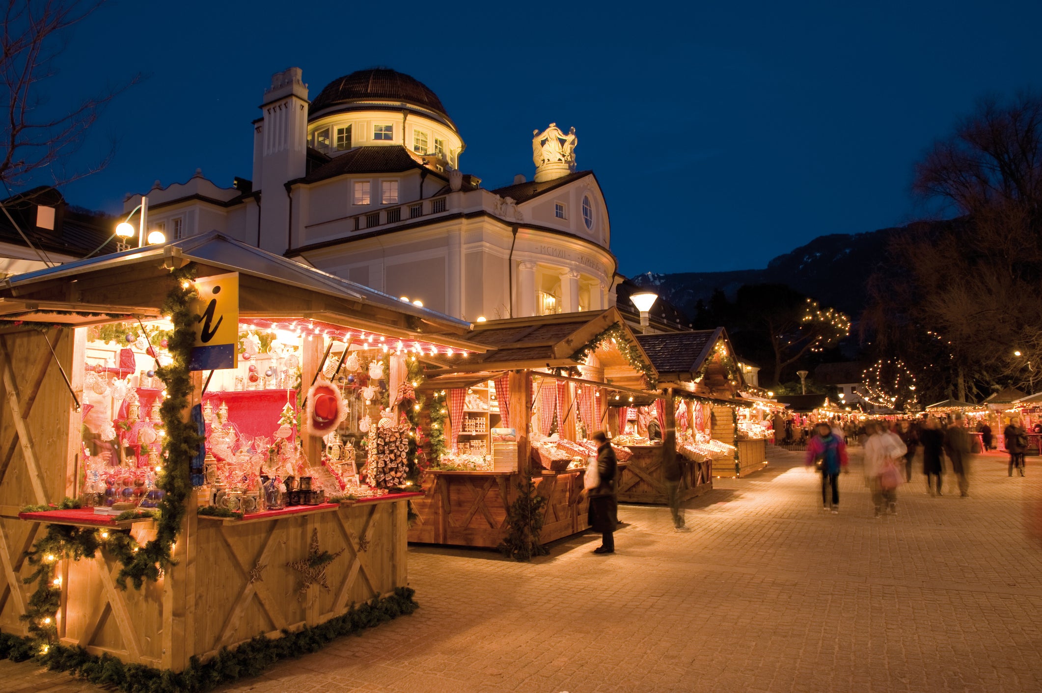 A festive outdoor holiday market at night with warmly lit wooden stalls, decorated with lights and garlands, and people browsing. A grand building is in the background under a dark blue sky.