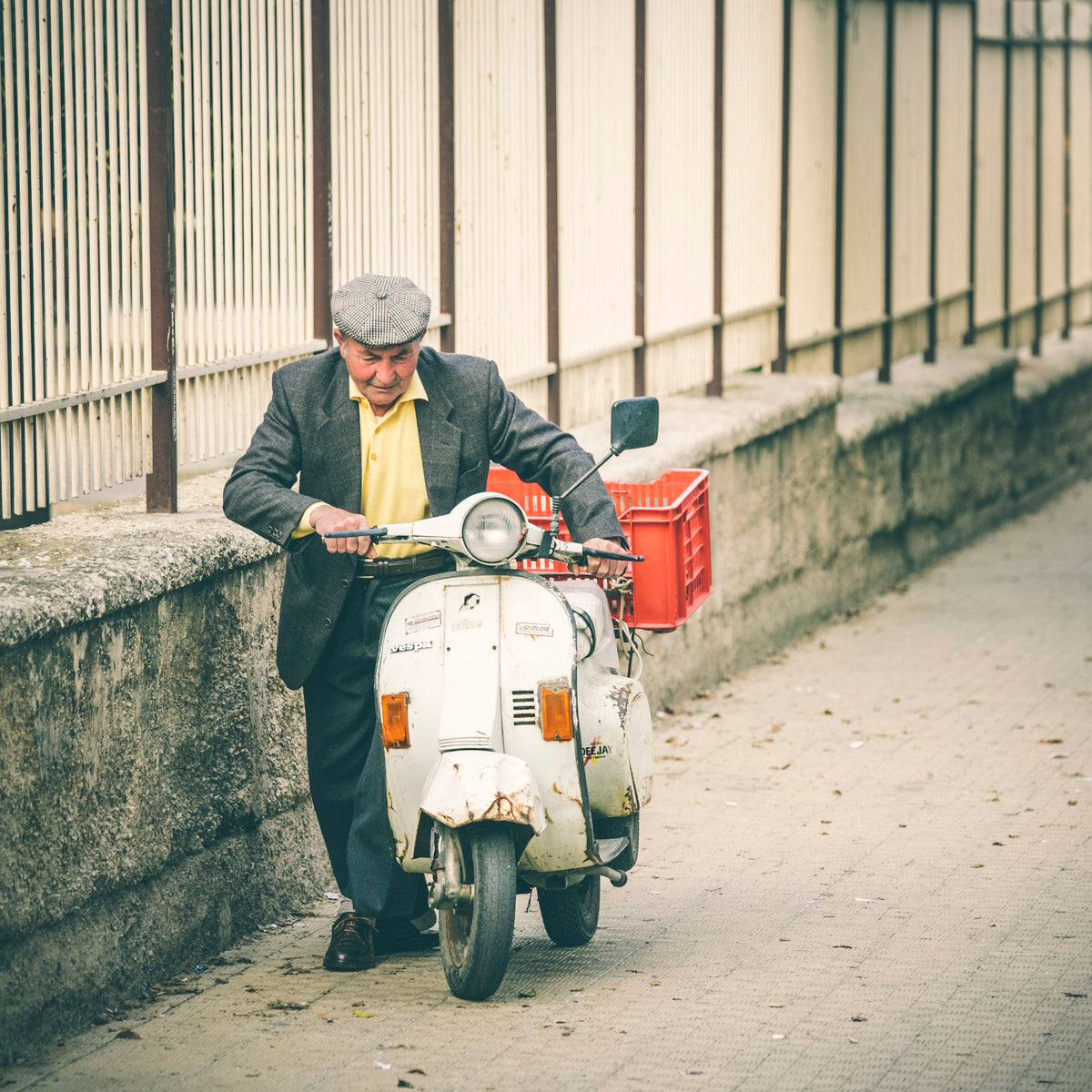 An elderly man in a flat cap and blazer walks his vintage scooter along a sidewalk next to a fence, with a red crate attached to the back of the scooter.