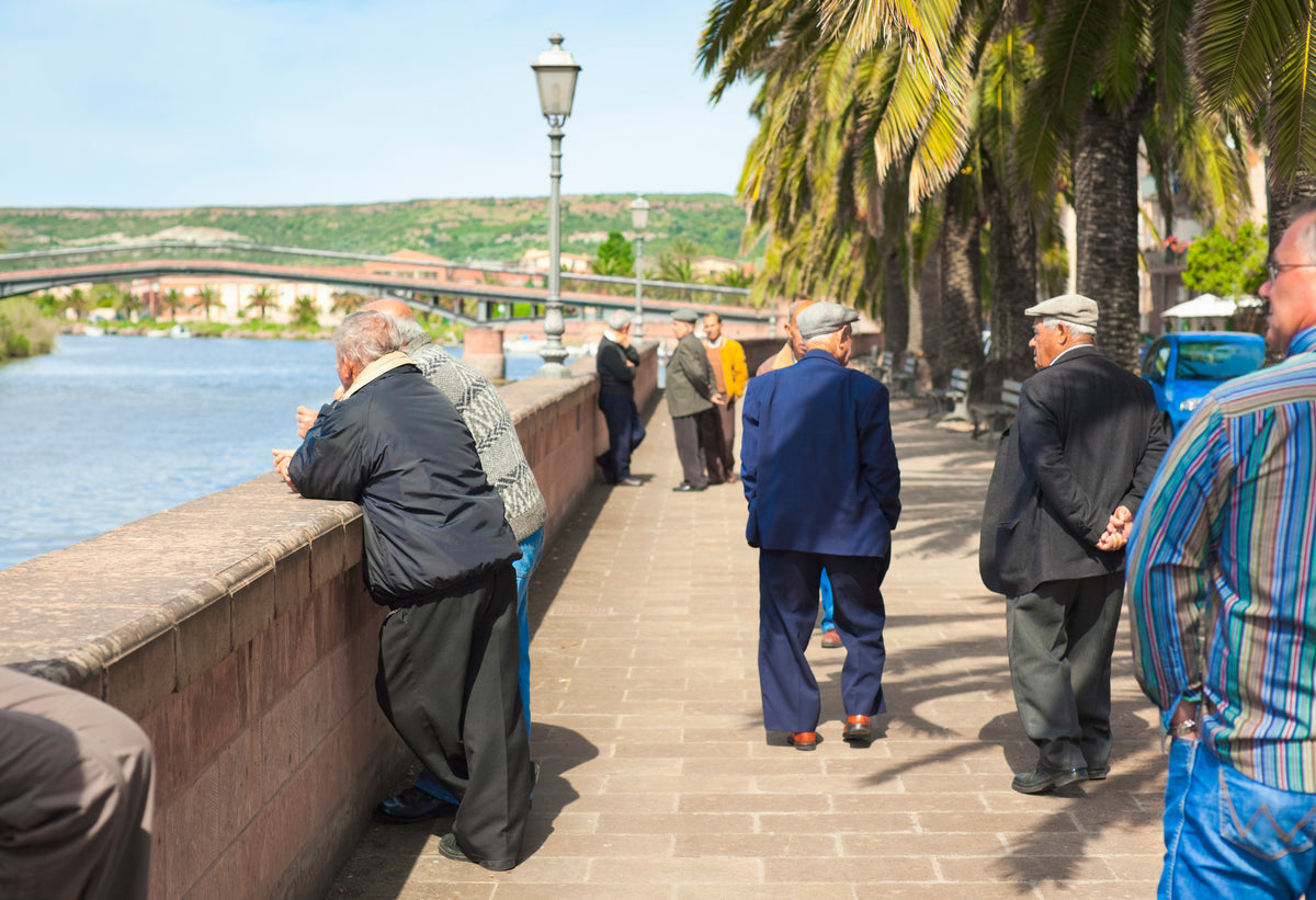 Elderly people walk and chat along a sunny riverside promenade lined with palm trees. Some lean on the stone wall, while others stroll in small groups.