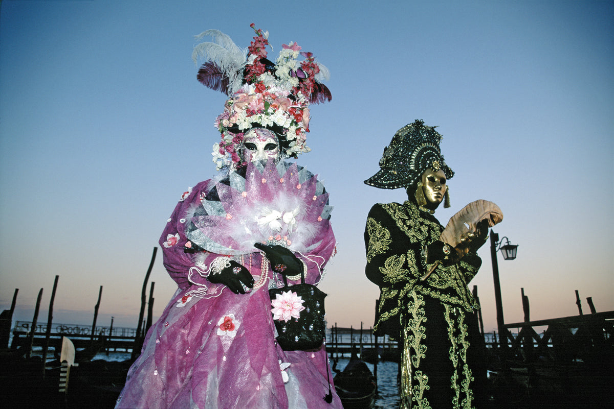 Two people in ornate costumes and masks stand outdoors at sunset; one wears a pink floral outfit with a large fan, the other wears a black and gold ensemble holding a mask, with wooden posts and water in the background.