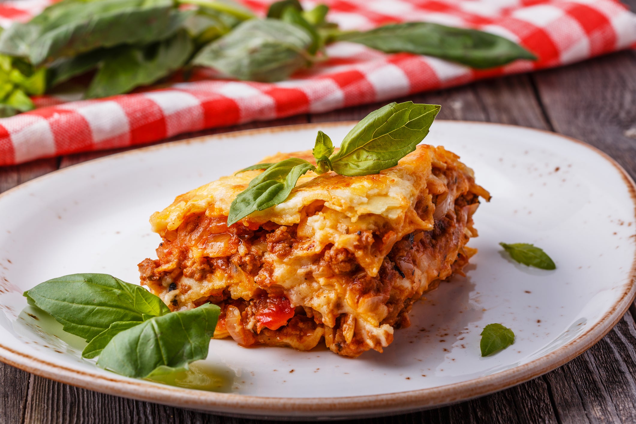 A slice of lasagna topped with fresh basil sits on a white plate, with more basil leaves around it. A red and white checkered cloth and basil leaves are in the background on a wooden table.