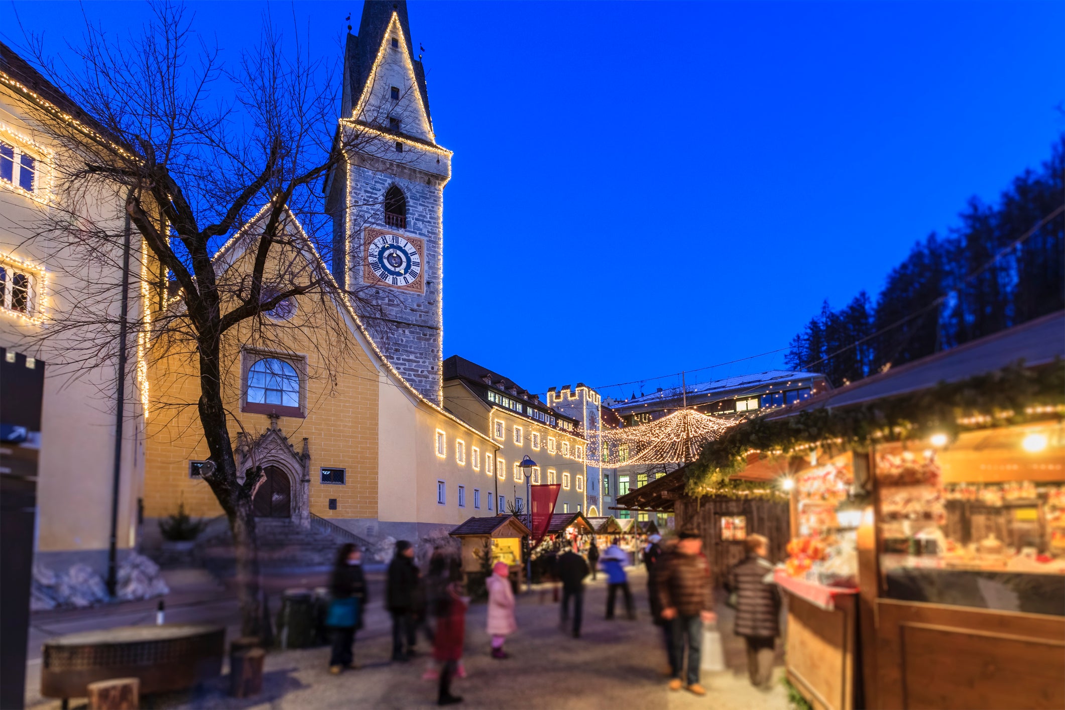 A festive outdoor market with blurred crowds, Christmas lights, and a tall clock tower at dusk. Stalls are decorated and glowing, creating a cheerful holiday atmosphere.