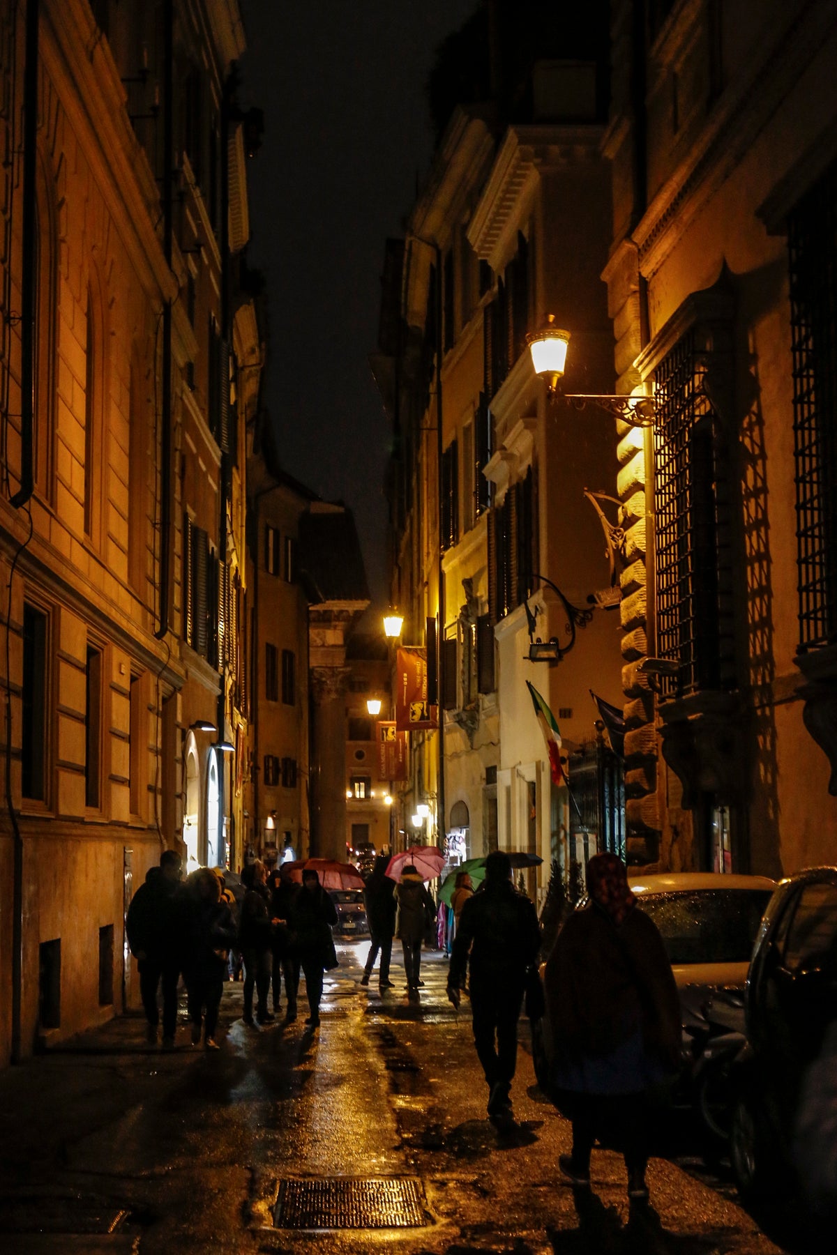 A narrow, dimly lit street at night with people walking along wet pavement, surrounded by tall buildings with warm lights and some parked cars.