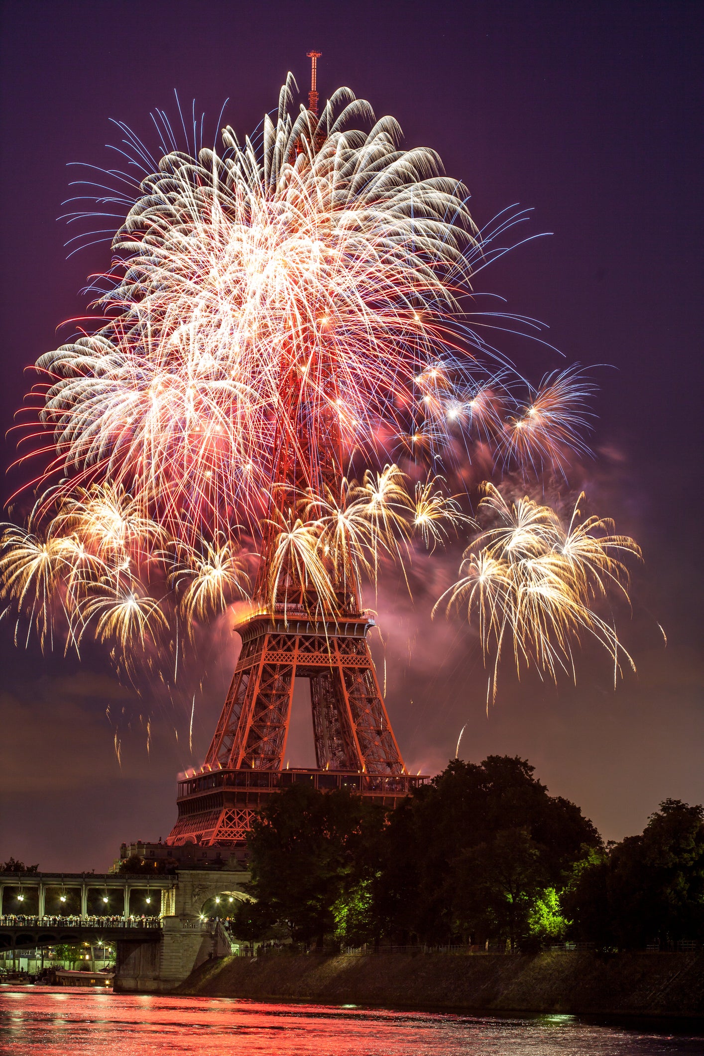 Fireworks explode in vibrant colors above the Eiffel Tower at night, reflecting on the river below, with trees silhouetted in the foreground.