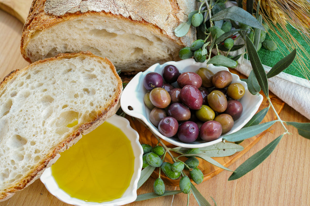 A sliced loaf of rustic bread, a bowl of olives, and a small dish of olive oil are arranged on a wooden board with olive branches and leaves.