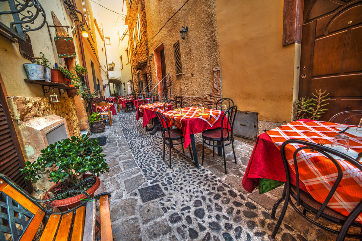 A narrow cobblestone street lined with small tables covered in red-checkered cloths, black chairs, potted plants, and warm yellow-toned buildings, evoking a cozy outdoor café atmosphere.
