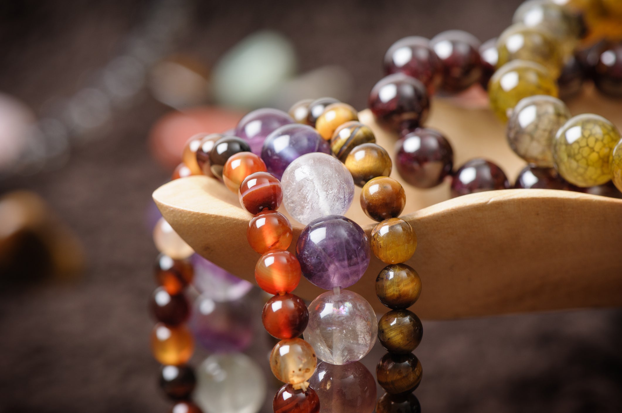 Close-up of colorful beaded gemstone bracelets draped over a wooden display, featuring beads in shades of purple, red, brown, and green, with a blurred dark background.