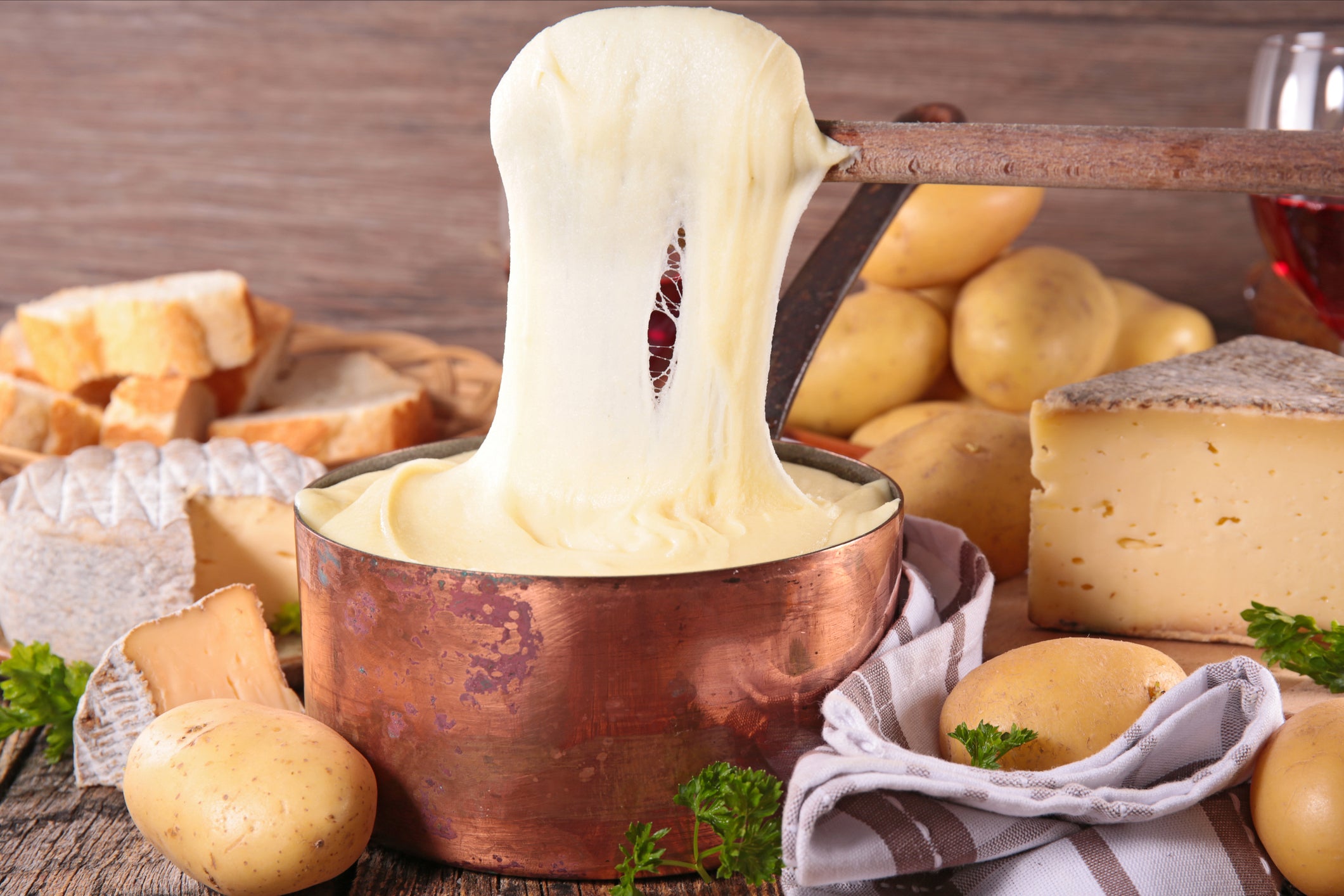 A pot of melted cheese is being stirred with a wooden spoon, surrounded by potatoes, bread, and cheese wedges on a rustic table.