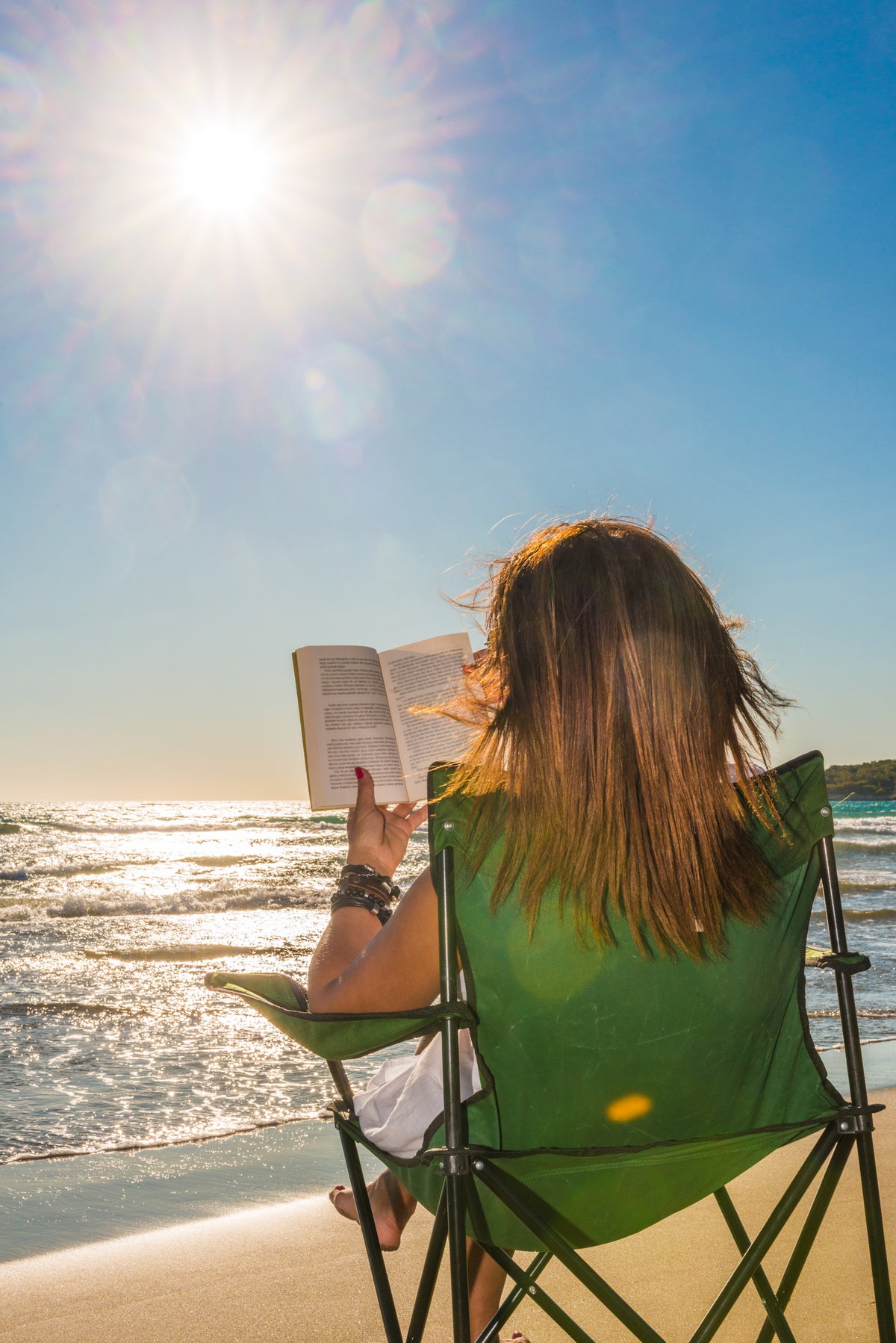 A person sits in a green chair on the beach, reading a book under the bright sun with waves in the background and a clear blue sky.