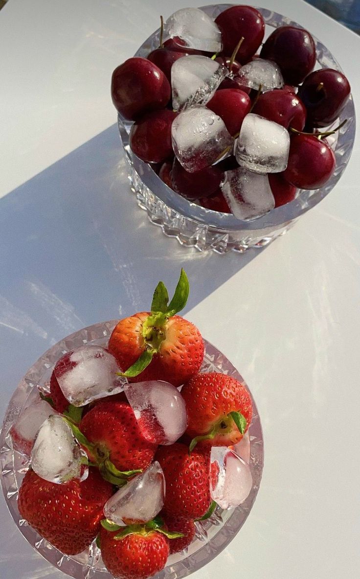 Two glass bowls on a white surface, one filled with fresh cherries and ice cubes, the other with strawberries and ice cubes, both bathed in natural light.