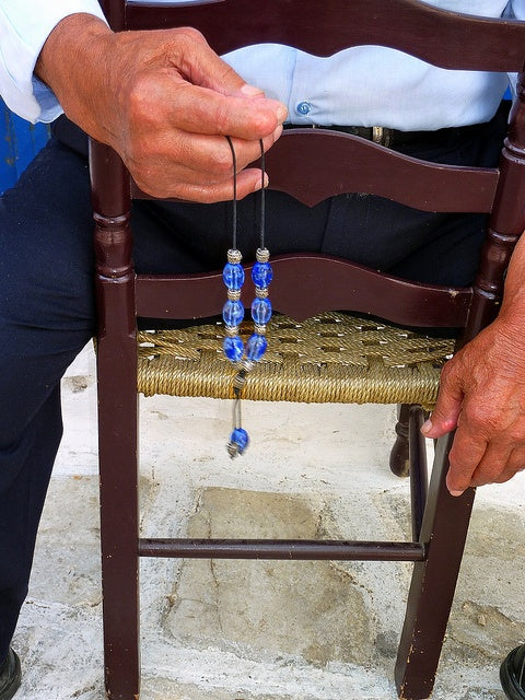 A person sitting on a woven chair holds a string of blue glass worry beads in their right hand, with their left hand resting on the chair. Only the persons torso and hands are visible.