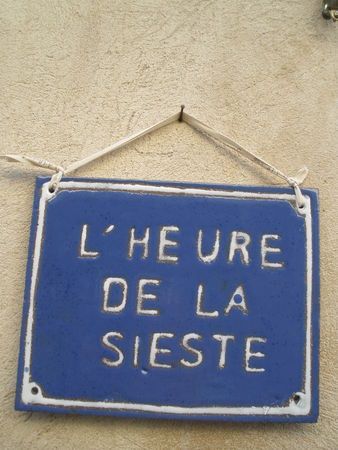 A blue rectangular sign with white letters reads LHEURE DE LA SIESTE, which is French for nap time. The sign is hanging by a string against a beige wall.