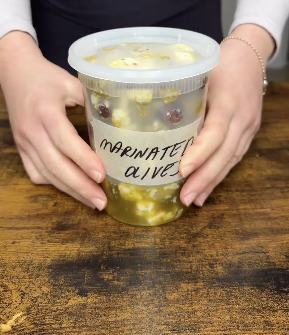 Hands holding a plastic container of marinated olives with feta cheese on a wooden surface.