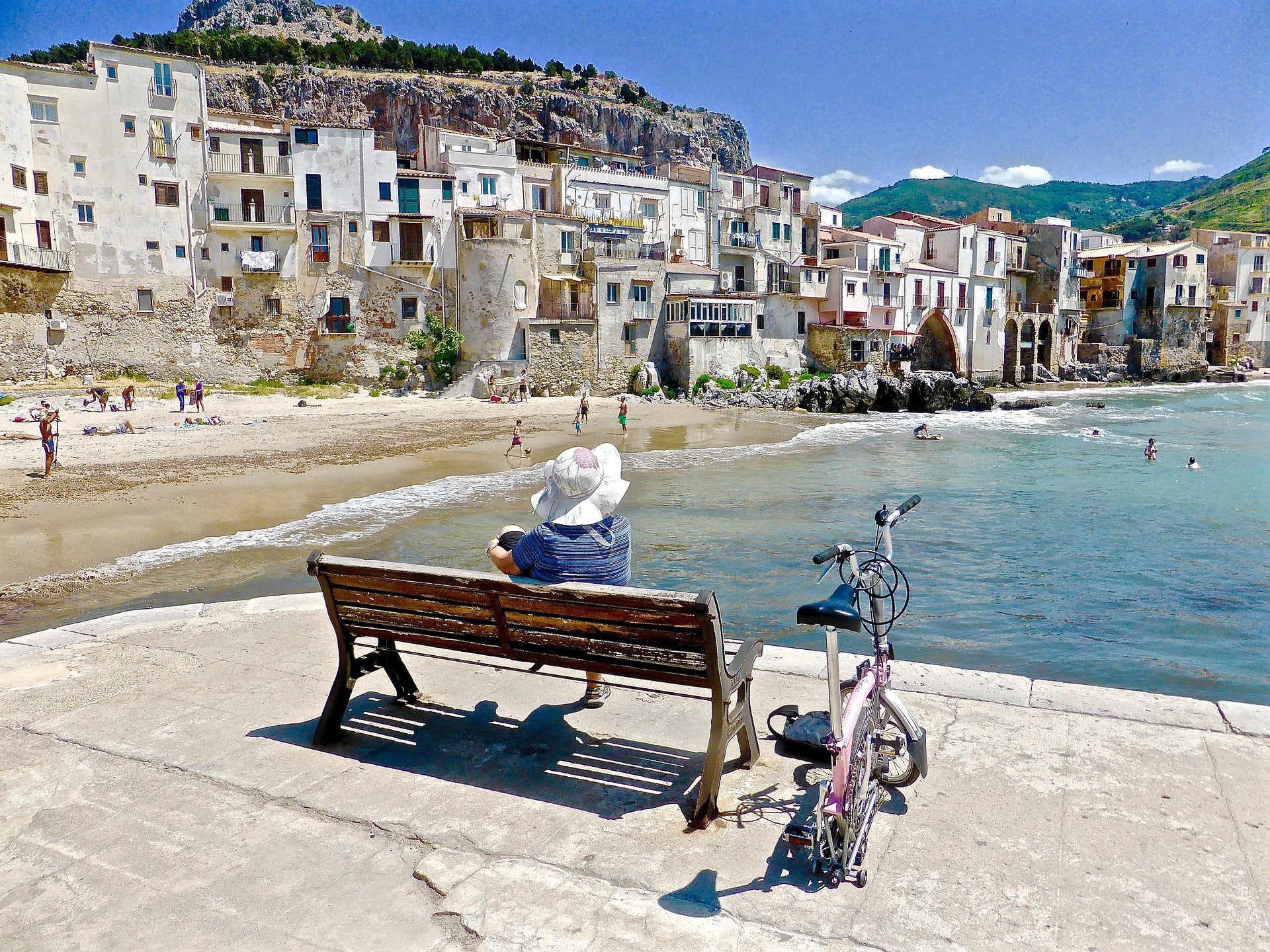 A person wearing a sunhat sits on a bench by the sea, with a bicycle nearby. People swim and play on a sandy beach with old buildings and hills in the background under a clear blue sky.