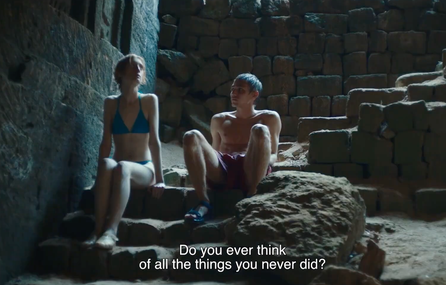 Two people in swimsuits sit on stone steps inside a cave-like structure, illuminated by soft sunlight. The scene has English subtitles: Do you ever think of all the things you never did?.